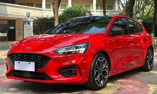 Красный автомобиль с чёрными колёсными дисками и решёткой радиатора. Вокруг деревья и здание с колоннами.