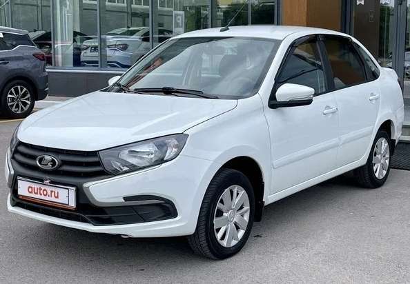 Белый автомобиль марки Lada стоит на улице возле автосалона. На номерном знаке написано «auto.ru». Машина имеет четыре двери и четыре колеса.