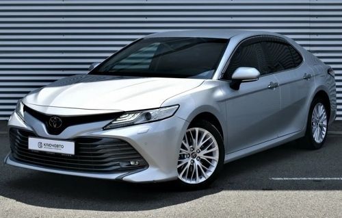 На изображении серебристый седан Toyota с хромированными элементами. Машина стоит на фоне серых гофрированных панелей.